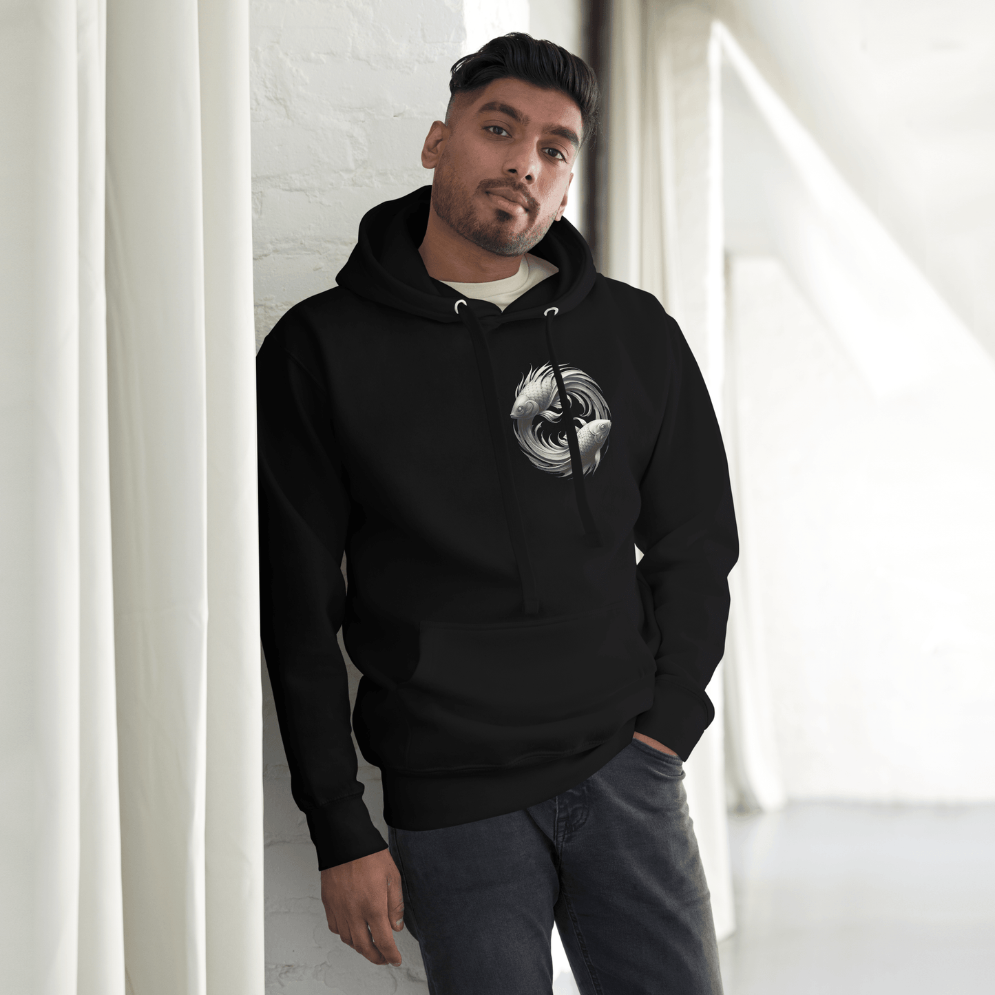 A man is leaning casually against a wall, wearing the black Deadpan Couture hoodie. The front of the hoodie displays a small, detailed design of two stylized fish in a circular pattern on the left chest, symbolizing the Pisces zodiac sign. The minimalist yet striking design complements the urban streetwear style.