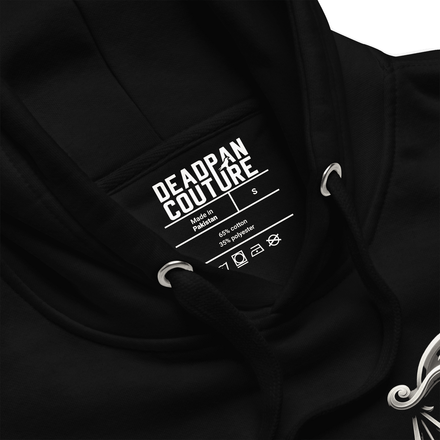 showing product details: A close-up view of the black Deadpan Couture hoodie, focusing on the inside label. The label displays the brand name, size (S), and fabric composition (65% cotton, 35% polyester). The image also includes a partial view of the ornate Libra scales design on the left chest, emphasizing the hoodie’s quality and attention to detail.